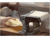 A hand is turning a crank of a pasta machine to get the dough through the roller. The other hand is holding the dough.